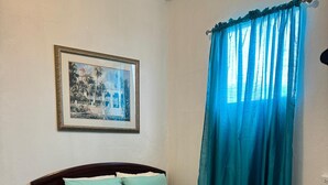 Una cama bien hecha con una colcha gris y dos almohadas, un cuadro en la pared y una ventana con cortinas azules.