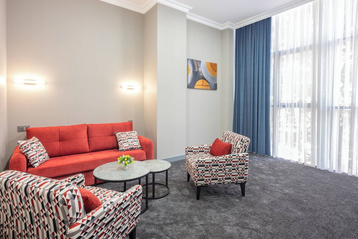 A modern hotel room with a red sofa, patterned armchairs, a small round table, and a large window with blue curtains.