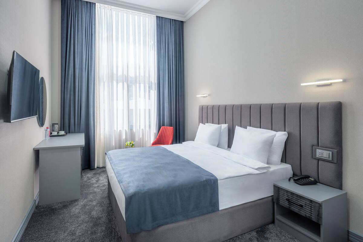 A modern hotel room with a large bed, a bedside table, a flat-screen TV, and a window with curtains.