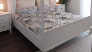Nelipylväinen sängynpäätyyn kiinnitetty katos, jossa on pehmustettu peitto, yöpöydät lampuineen ja ruukkukasvi.