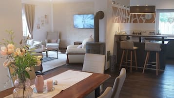Un salon moderne avec une table à manger en bois, des sièges confortables, un grand luminaire suspendu et un coin cuisine équipé d’une cuisinière et de tabourets de bar.