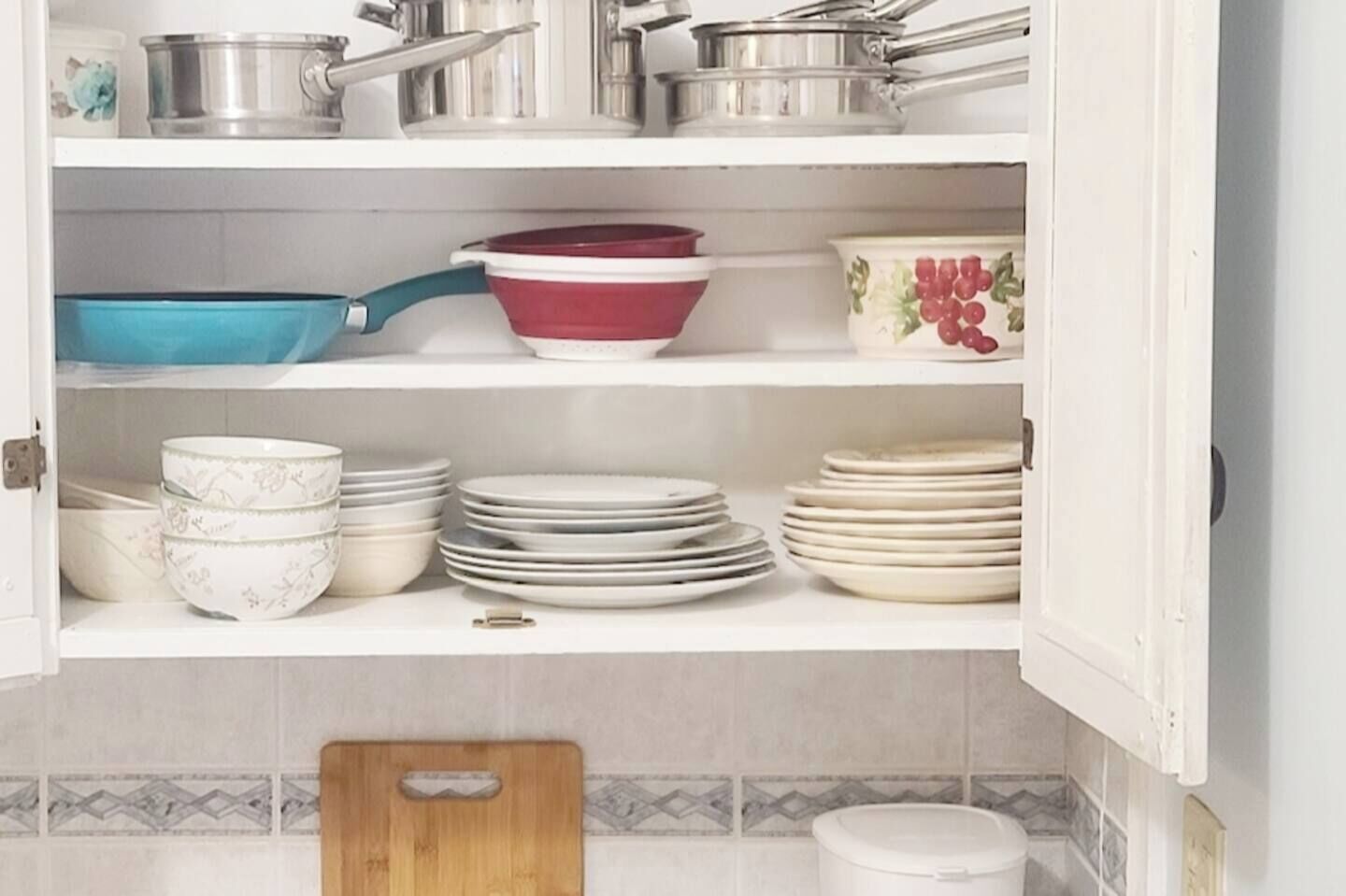 Una cocina con armarios blancos, una tabla de cortar de madera y diversos utensilios de cocina, incluyendo ollas, sartenes y platos.