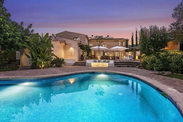 A swimming pool with a stone deck, a patio area with outdoor seating, and a building with a stone facade.