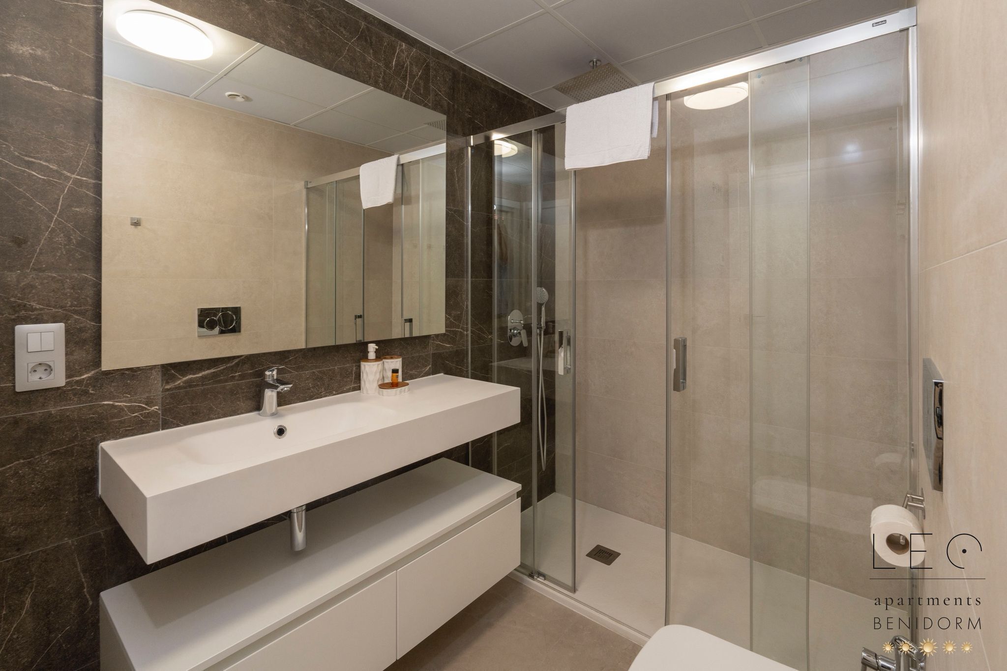 Un baño moderno con pared de mármol, tocador blanco, cabina de ducha de vidrio y dispensador de papel higiénico.