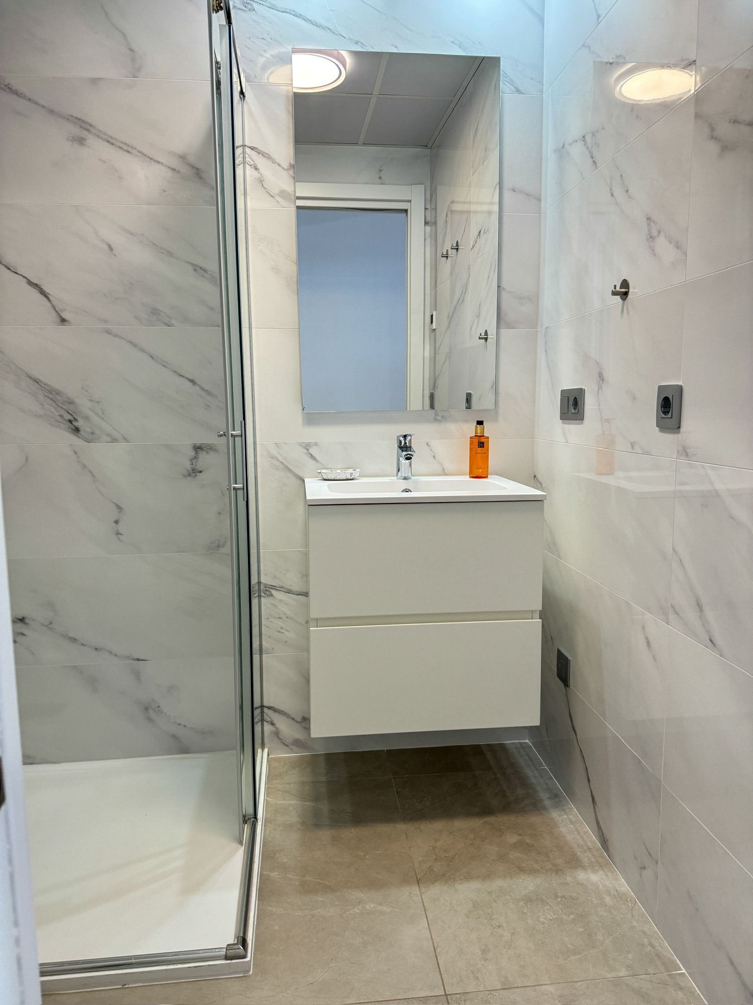 A modern bathroom with a glass shower enclosure, a white vanity with a mirror, and marble walls.