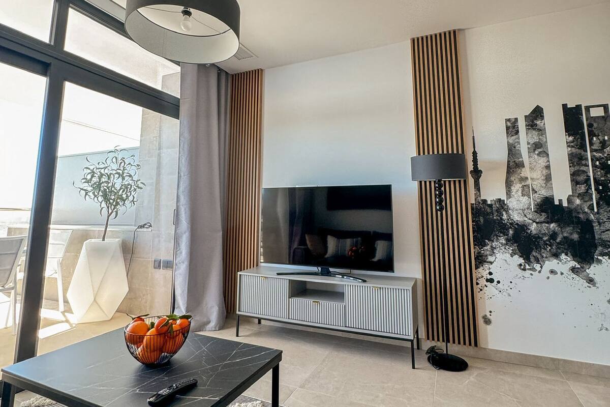 A modern living room with a TV, a dining table with a bowl of fruit, and a large window.