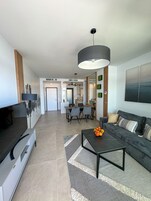 Una sala de estar moderna con sofá, mesa de centro y televisor. Se observa un comedor con sillas y una mesa al fondo.