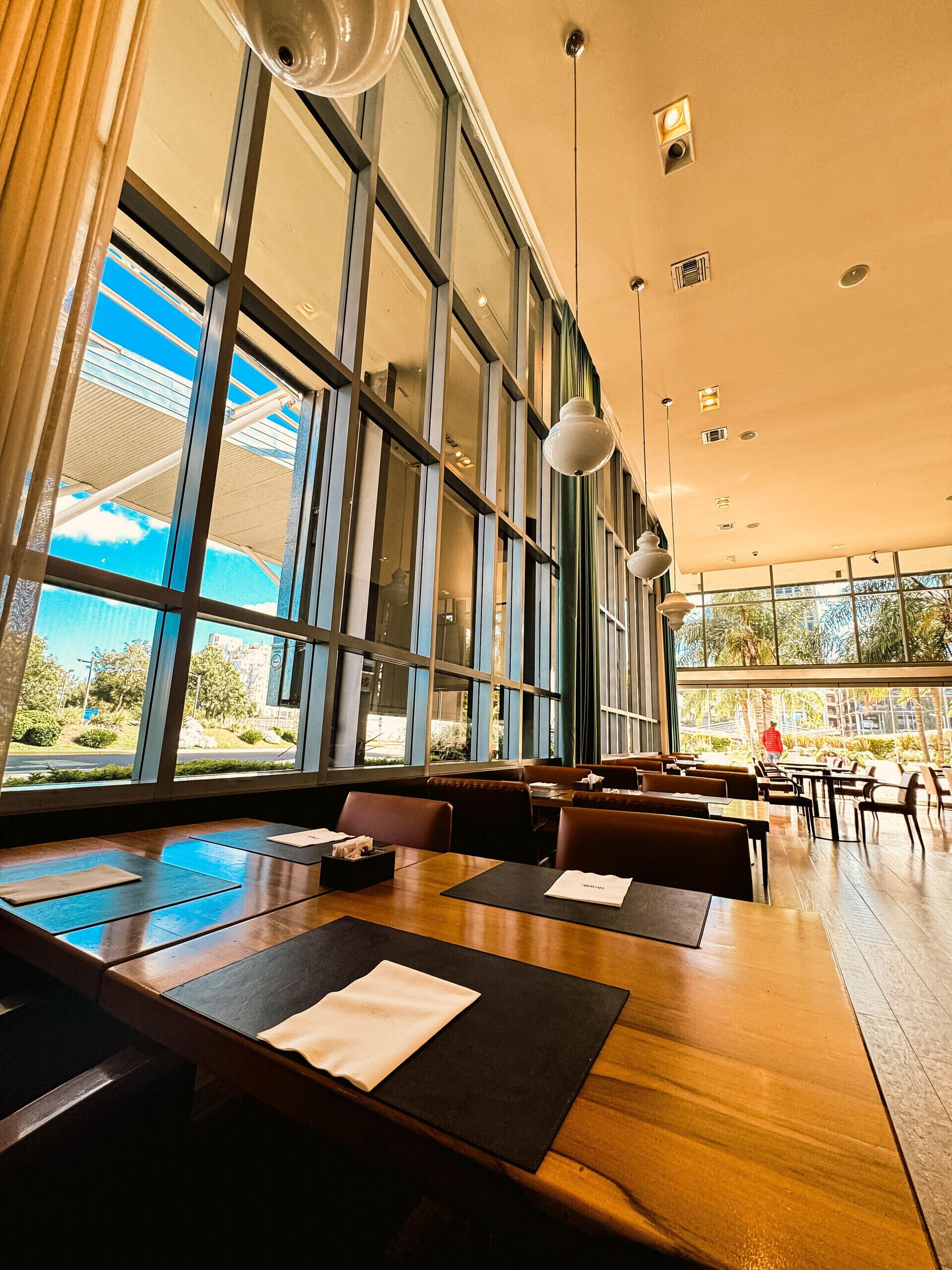 Un restaurante espacioso con amplios ventanales, mesas de madera e iluminación colgante moderna.