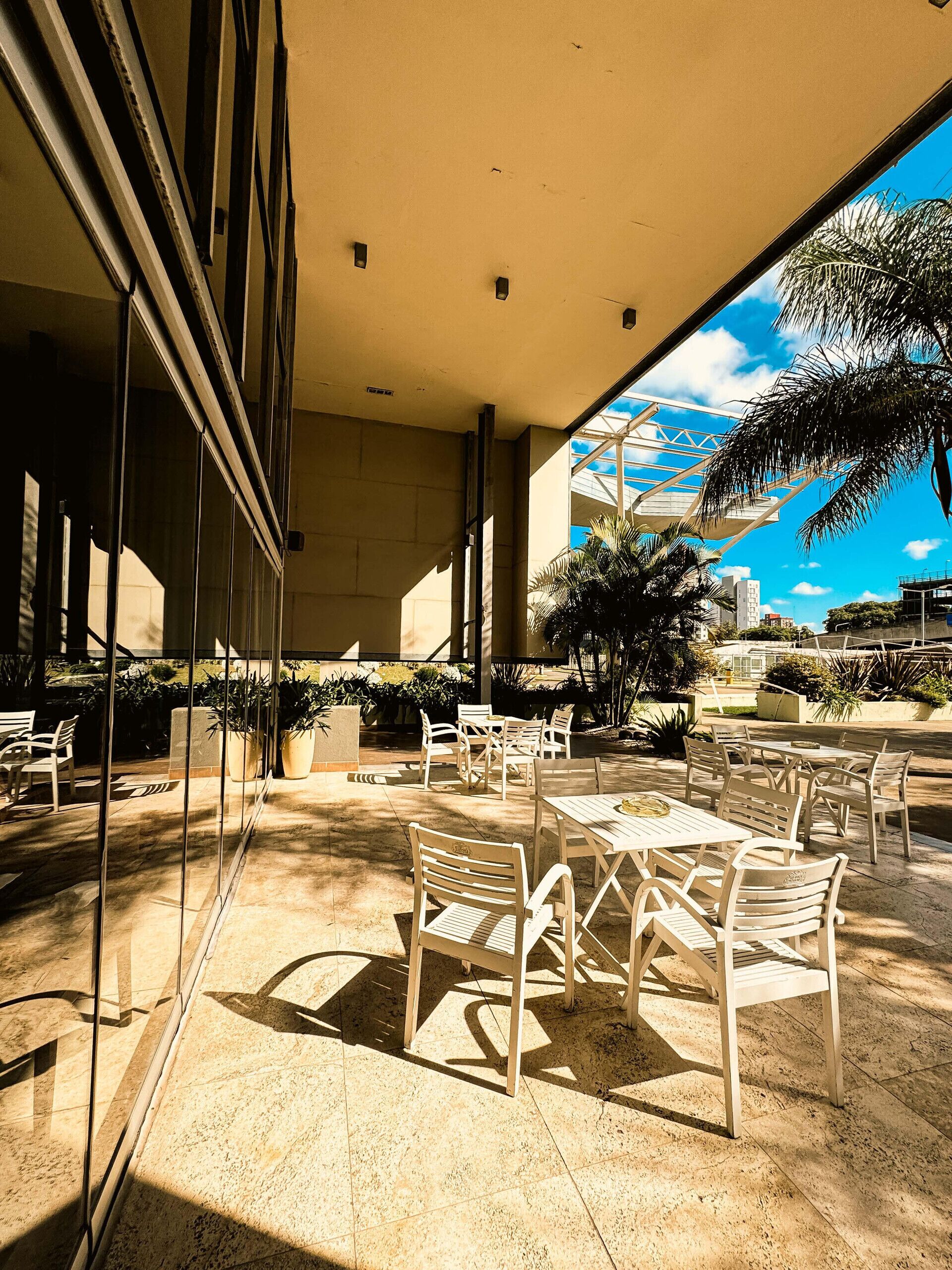 Un patio al aire libre con mesas y sillas, ventanas amplias y vistas a palmeras y edificios.