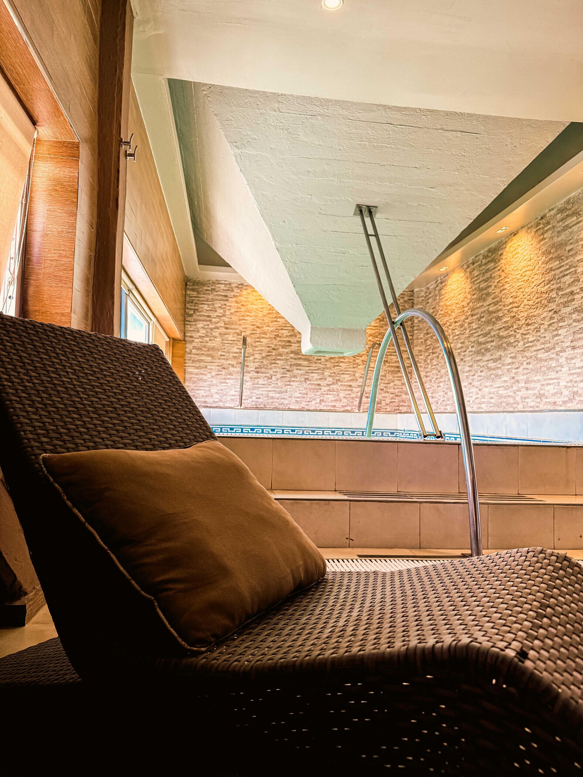 Una habitación con una silla de mimbre, un cojín marrón, una escalera metálica y una pared de ladrillo.