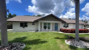 A single-story house with a front porch, a blue door, and a small front yard with red flowers and a tree. - Harbor Side Retreat (Port Charlotte)