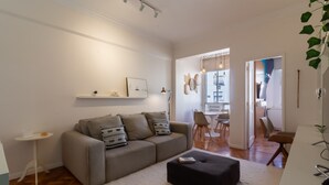 A modern living room with a sofa, ottoman, and a small side table. There is a dining area visible through an open doorway.