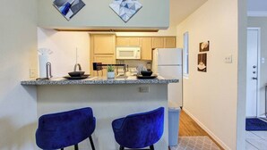 A compact kitchen with two blue bar stools, a microwave, a refrigerator, and a small island with a sink. - HTX Cozy Hideaway (Houston)