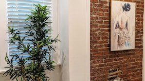 Une pièce avec un plant de bambou dans un pot noir, une fenêtre avec des stores, un mur en briques et un tableau.