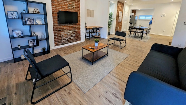 Un salon moderne avec un mur d’accent en briques, un téléviseur à écran plat, des chaises en métal noir, une table basse et un coin repas avec une table et des chaises.