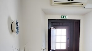 Une pièce avec une porte en bois foncé, un mur blanc, un sol carrelé, un vase avec des branches et un appareil de climatisation.