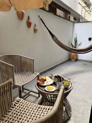 A wicker chair and table set up on a balcony with a hammock, a potted plant, and a breakfast spread including toast, eggs, and fruit.