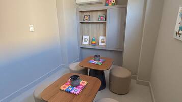 A small, well-lit room with two wooden tables, each with a colorful puzzle mat and a black mug. There are four beige stools around the tables, and a gray cabinet with shelves against the wall.