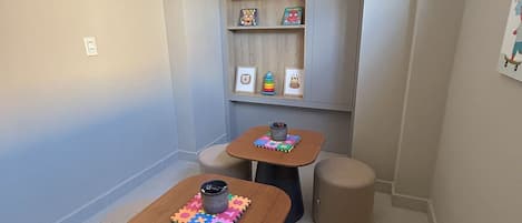 A small, well-lit room with two wooden tables, each with a colorful puzzle mat and a black mug. There are four beige stools around the tables, and a gray cabinet with shelves against the wall.