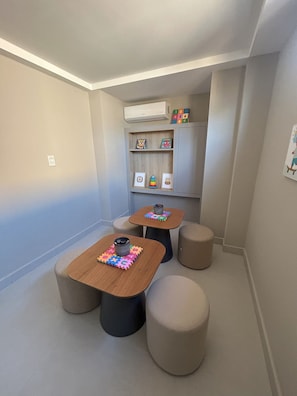 A small, well-lit room with two wooden tables, each with a colorful puzzle mat and a black mug. There are four beige stools around the tables, and a gray cabinet with shelves against the wall. - Flat Cabo Branco Aurea 50m do mar 105 (Joao Pessoa)