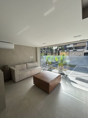A modern living room with a beige sofa, a brown ottoman, and a glass coffee table. - Flat Cabo Branco Aurea 50m do mar 105 (Joao Pessoa)