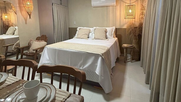 A hotel room with a bed, a dining table with plates, a chair, a small table with a basket, and a wall-mounted air conditioner. - Flat Cabo Branco Aurea 50m do mar 105 (Joao Pessoa)