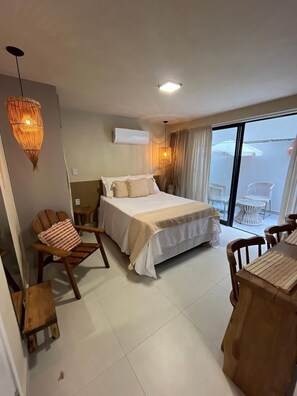 A bedroom with a bed, a wooden chair, a small table, and a balcony with chairs. - Flat Cabo Branco Aurea 50m do mar 105 (Joao Pessoa)