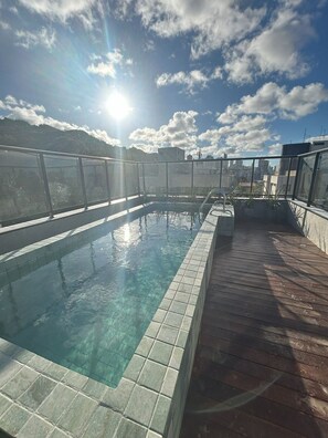 A rooftop pool with clear water, surrounded by a wooden deck and a glass railing.