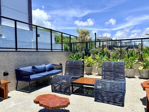 An outdoor seating area with a sofa, chairs, and a wooden table, surrounded by potted plants and a cityscape view. - Flat Cabo Branco Aurea 50m do mar 105 (Joao Pessoa)