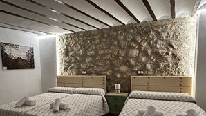 Ein Zimmer mit zwei Betten, jedes mit einem gemusterten Bettbezug und Handtüchern, an einer Wand aus strukturiertem Stein. Zwischen den Betten steht ein Nachttisch mit einer Lampe, und an der Wand hängt ein gerahmtes Bild.