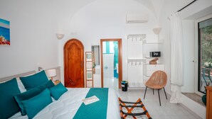 A bedroom with a bed, a wooden door, a white wall, a white ceiling, a white floor, a white chair, a white table, a white shelf, a white curtain, a white window, and a white radiator. - Room-breathtaking Suite With Sea View in (Positano)