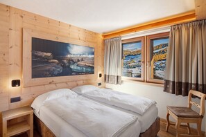 Una stanza in legno con un letto, una sedia, un quadro e una finestra con tende.