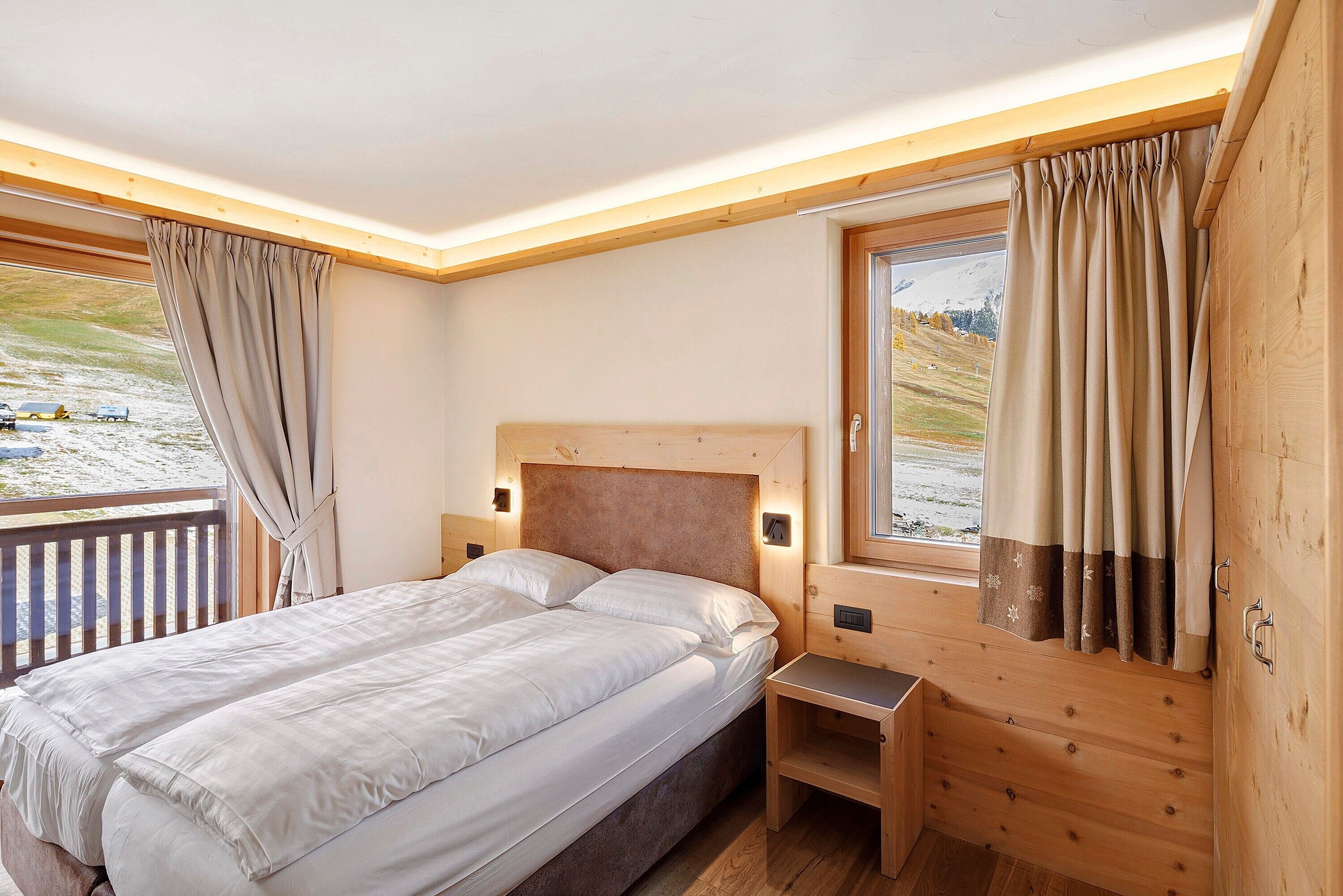 A bedroom with a wooden headboard, a bedside table, a window with curtains, and a view of a snowy landscape.