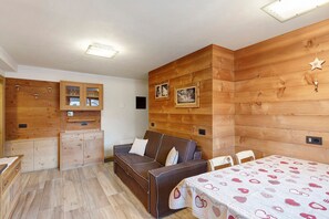 A cozy room with a wooden wall, a sofa, a table with a patterned tablecloth, and a cabinet with glass doors. - Apartment 'Gembro' with Mountain View, Shared Garden and Wi-Fi (Livigno)