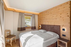 A bedroom with a wooden headboard, two beds, a nightstand, a chair, and a window with curtains. - Apartment 'Gembro' with Mountain View, Shared Garden and Wi-Fi (Livigno)