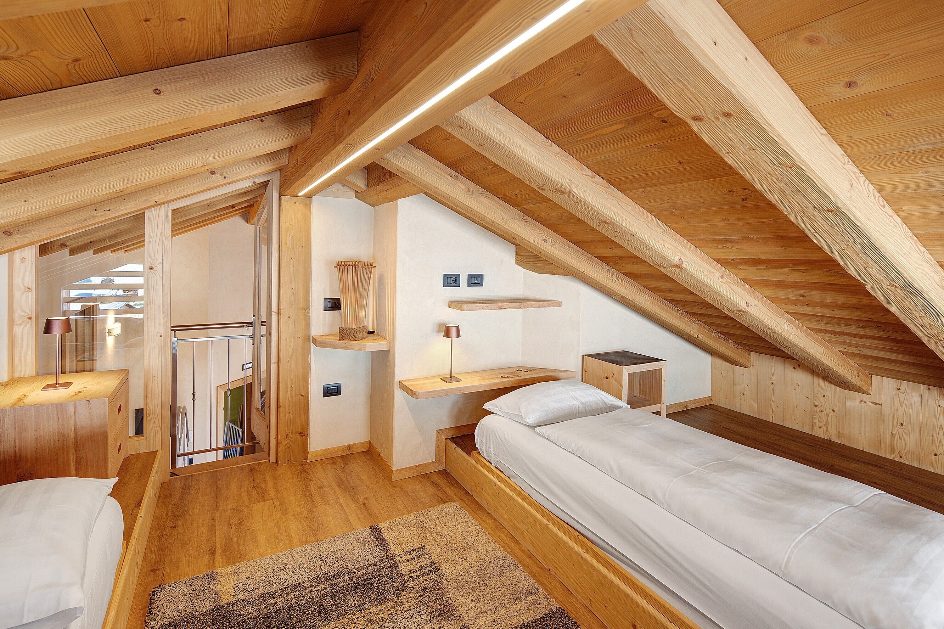 A loft bedroom with a wooden ceiling, a bed, a nightstand, and a small desk with a lamp.