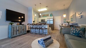 A modern living room with a large flat-screen TV, a round coffee table with towels, a gray sofa with decorative pillows, a kitchen area with a refrigerator and microwave, and a dining area with bar stools. - 4 BRw/WD | Dog Friendly | Balcony | Pool | Hot Tub (Corpus Christi)