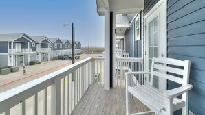 A balcony with a white bench, overlooking a row of beach houses with a clear sky. - 4 BRw/WD | Dog Friendly | Balcony | Pool | Hot Tub (Corpus Christi)