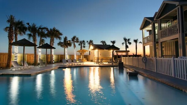 A swimming pool with lounge chairs and palm trees, surrounded by a wooden fence and a building with balconies. - 4 BRw/WD | Dog Friendly | Balcony | Pool | Hot Tub (Corpus Christi)