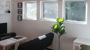 A modern living room with a black sofa, a small white table, a floor lamp, and a potted plant. There are three windows providing natural light.