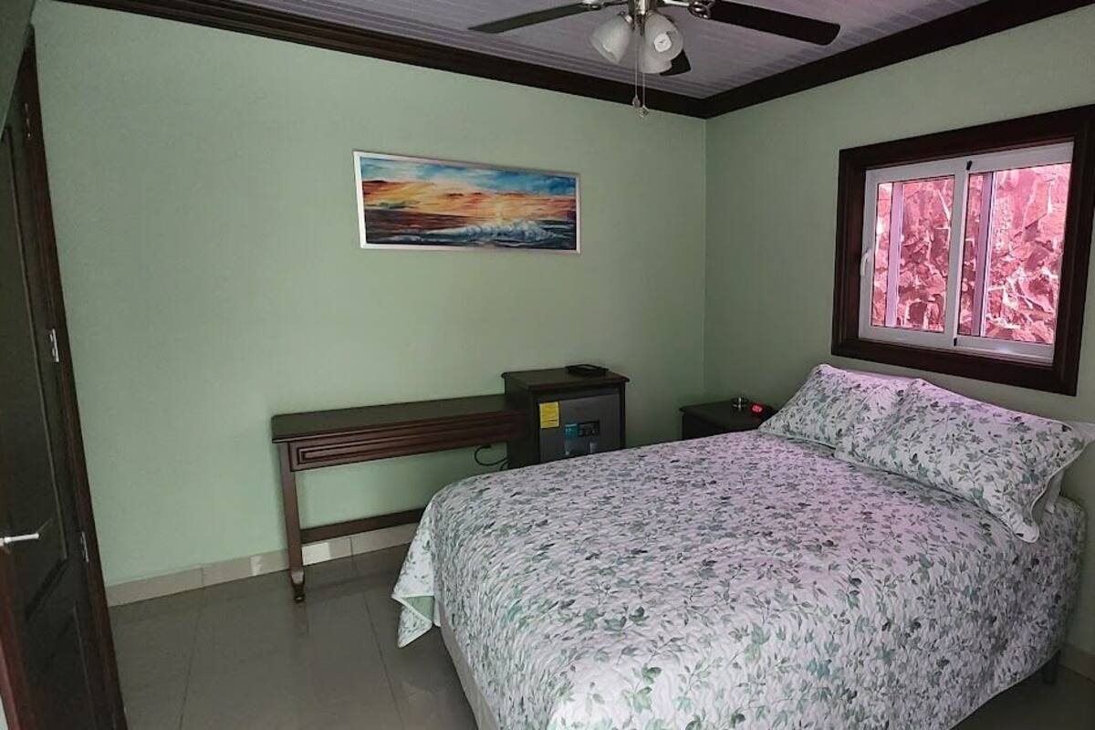 A bedroom with a bed, a nightstand, a dresser, a painting, and a window with a view.