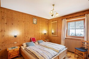 Une pièce aux murs lambrissés, comprenant un lit, des tables de chevet, une chaise, une fenêtre avec des rideaux et un luminaire fixé au mur.