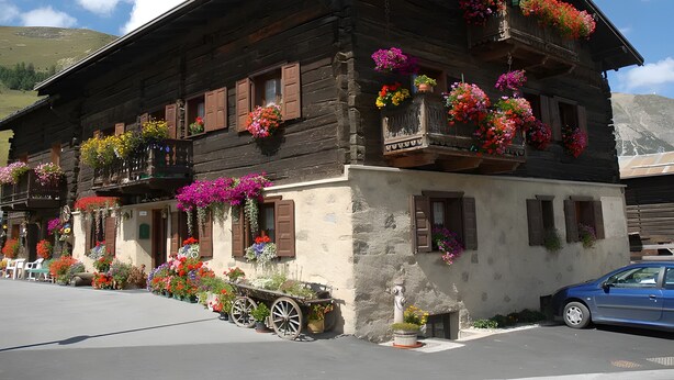 A wooden building with balconies adorned with flowers, a cart, and a car parked in front. - Apartment 'Di Bernardin' with Mountain View and Wi-Fi (Livigno)