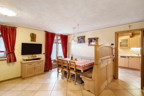 A compact room with a dining table, chairs, a television, a wooden bench, and a kitchen area.