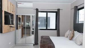 Un dormitorio moderno con cama, televisor montado en un panel de madera, puerta corredera de cristal y ventana con cortinas.