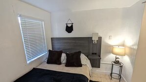 A bedroom with a bed, a bedside table with a lamp, a window with blinds, and a wall-mounted shelf. - Bear cave! (Tucson)