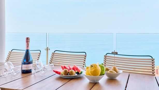 Een tafel met een fles wijn, glazen en schalen vol fruit, bij een raam met uitzicht op de zee.