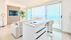 Een moderne woonkamer met een bank, een salontafel met een ingebouwd inductiefornuis, een aan een houten paneel bevestigde televisie en een balkon met uitzicht op zee.
