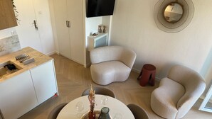 A modern living room with a round dining table, two beige armchairs, a small red stool, a TV mounted on the wall, and a decorative mirror. - Waterfront apartment (sea view) (Concarneau)