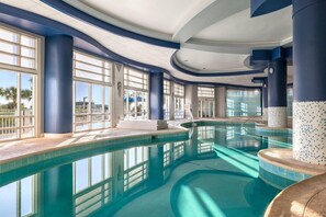 Una amplia piscina interior con agua cristalina y azul, rodeada de grandes ventanales y pilares azules.